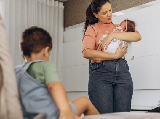 A mother lovingly cradles her baby while her child sits nearby on the couch in a modern room setting.