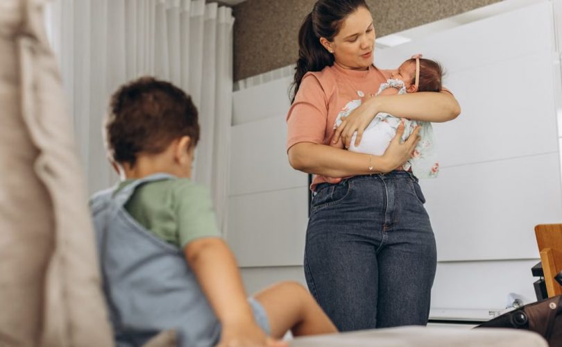 A mother lovingly cradles her baby while her child sits nearby on the couch in a modern room setting.