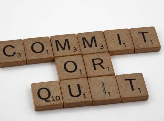 Wooden Scrabble tiles arranged to form the words 'Commit or Quit' on a white background.